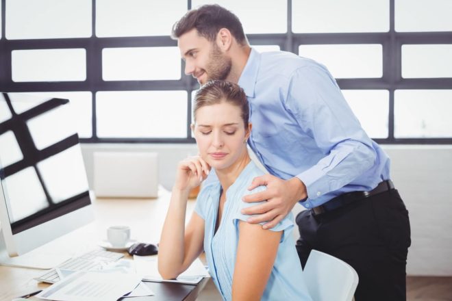 Mann legt Hand auf Schulter der vor ihm am Computer sitzenden Frau. Belästigung am Arbeitsplatz ist der Mitarbeiterzufriedenheit nicht zuträglich