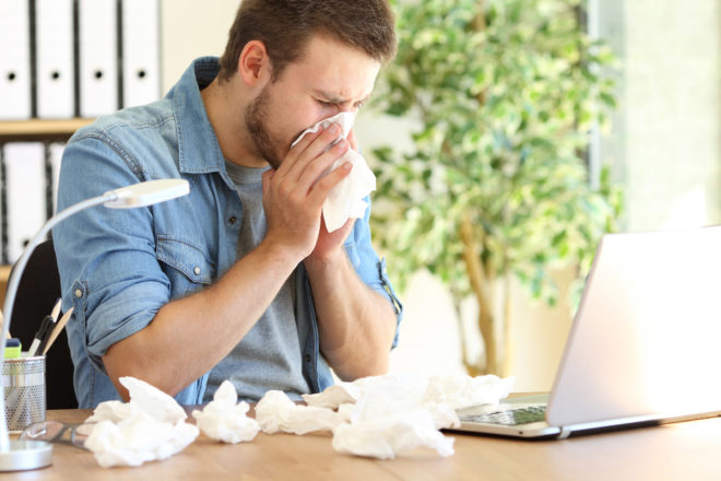 Foto zeigt einen sich schnäuzenden Mann mit benutzten Taschentüchern vor sich und seinem Laptop zum Artikel über Lohnfortzahlung bei Krankheit