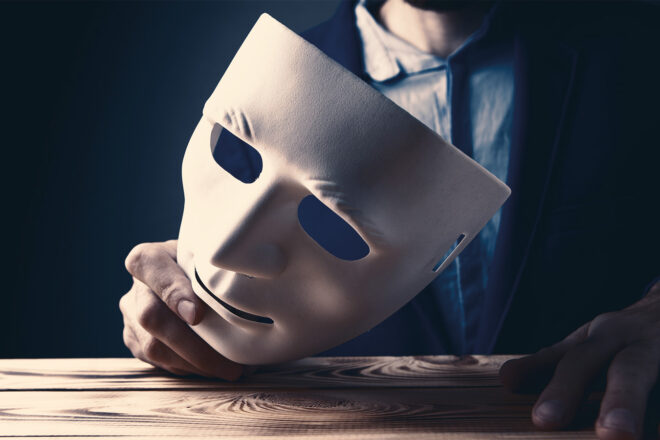 Symbolbild Hinweisgeberschutzgesetz- Eine Person sitzt im dunklen Hintergrund des Bildes und hält eine weiße Maske in der Hand