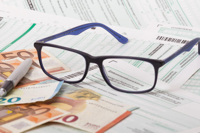 Foto zeigt Brille auf Euroscheinen neben einem Kugelschreiber und darunter Formularen für die Einkommensteuer: selbstständig Tätige müssen hier ganz genau hinschauen