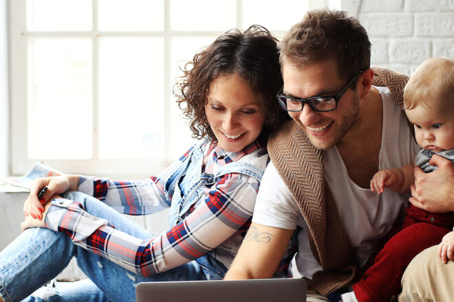 Foto zeigt eine Frau und ein Mann mit Baby auf dem Schoß, die auf dem Laptop Informationen über Elterngeld für Selbstständige suchen