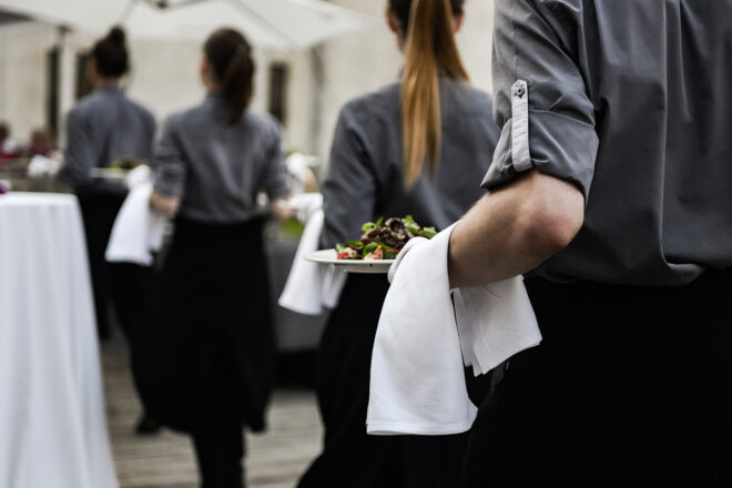 Foto zeigte eine Reihe von Servicekräften mit Tellern auf der Hand und Serviette über dem Arm, die im Ferienjob den Mindestlohn erhalten