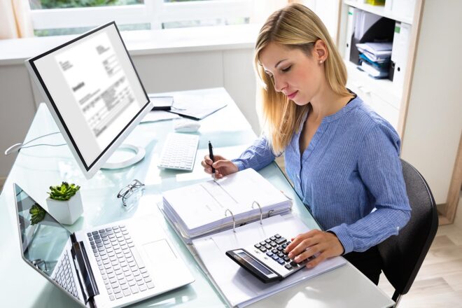 Foto zeigt Frau im Büro mit Taschenrechner und Ordner vor Monitor und Laptop die GoBD stets beachtend