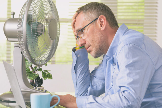 Ein schwitzender Mann am Schreibtisch symbilisiert, dass viele Arbeitnehmer bei einer Maximaltemperatur am Arbeitsplatz im Büro oder anderswo im Unternehmen gerne Hitzefrei von der Arbeit hätten
