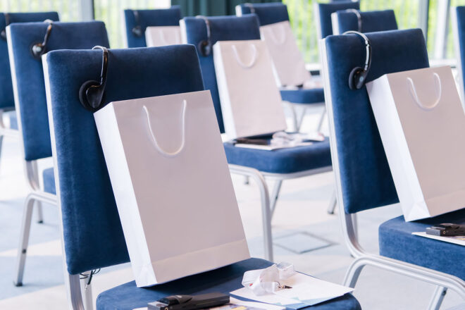 Das Bild von Stühlen in einem Veranstaltungssaal, auf denen Geschenktüten stehen, symbolisiert die Herausforderung für Unternehmen, Ideen für nützliche oder langfristig beliebte Werbegeschenke für Kunden zu finden und die Werbeschenke steuerfrei zu gestalten.