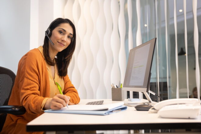 Symbolbild Weiterbildung: Frau mit orangener Bluse und Strickjacke sitzt am PC-Arbeitsplatz und macht sich Notizen